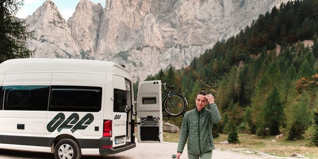 Ein VW Grand California Van steht vor einer Bergkulisse, eine Frau geht auf die Kamera zu.