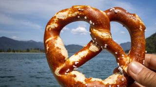 Brotzeit-Pause beim Campen in der Bayerischen Karibik am Campingplatz Walchensee: Eine Person hält vor dem See und Bergen im Hintergrund eine Breze in die Kamera