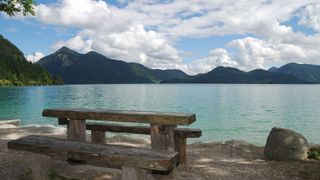 Der perfekte Picknickplatz beim Walchensee Camping: Ein Holztisch und Holzbänke am Ufer des Walchensees, im Hintergrund Berge und wolkiger Himmel 