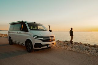 Mann steht bei Sonnenuntergang am Meer, neben ihm steht ein VW California Beach Camper.