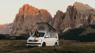 Mann sitzt auf Motorhaube eines VW Beach mit aufgestelltem Dachzelt. Hinter ihm ist eine imposante Berglandschaft zu sehen. 