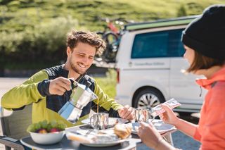 Ein Mann mit grüner Sportjacke schenkt sich beim Camping Trip mit Fahrrad am Campingtisch Kaffee in seine Tasse und lacht dabei, gegenüber sitzt eine Frau, die sich Marmelade auf ihr Brot schmiert, im Hintergrund ein Camper mit Fahrrädern