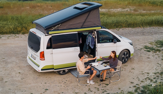 Pärchen sitzt vor dem Camper auf Campingstühlen in der Natur