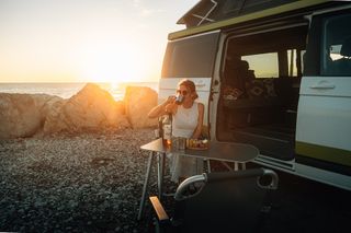 Eine Frau sitzt an einem Ufer vor einem offenen CamperBoys Camper am Campingtisch und trinkt aus einer Tasse.