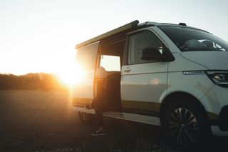 Ein VW California Van im Sonnenschein. Eine Frau sitzt in der geöffneten Schiebetür und bindet sich die Schuhe.