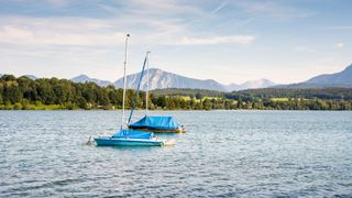 Zwei blaue Boote schwimmen auf dem Riegsee vor der Bergkulisse und dem von Bäumen gesäumten Ufer auf dem Wasser