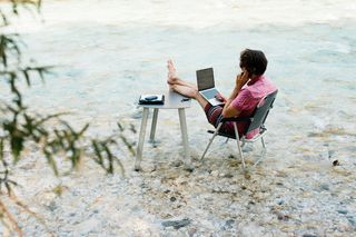 Ein Campingtisch und -stuhl sind in einem Bach aufgebaut. Ein Mann sitzt auf dem Stuhl, hat die Füße auf den Tisch gelegt, telefoniert und hat einen Laptop auf dem Schoß.