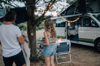 Ein VW Ocean Camper steht geparkt und ist mit einer Lichterkette geschmückt. Neben dem Van sind ein Campingtisch und -stühle aufgebaut, ein Mann und seine Tochter hängen Wäsche ab.