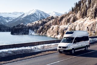 VW Grand California fährt über eine Brücke Im Hintergrund ist eine Winterlandschaft zu erkennen 