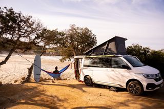 Woman with surfboard next to VW California Ocean on beach in hammock