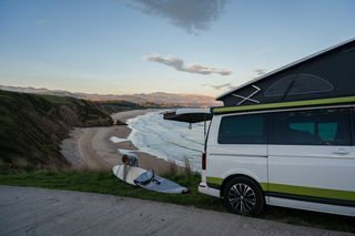 Urlaub im April an der Algarve: Eine Frau mit Surfboard neben ihrem Camper vor der Küste und Berglandschaft Portugals