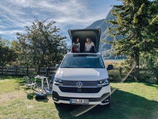 Zwei Frauen sitzen im geöffneten Aufstelldach eines VW-Busses in der Natur.