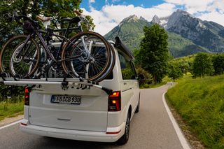 Fahrrad Camper auf einer Straße in den Bergen mit beladenem Fahrradträger von hinten fotografiert, während er vorbei an sattgrünen Wiesen fährt 