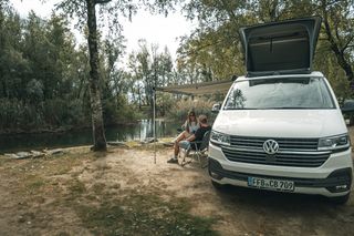 Zwei Menschen sitzen vor einem Camper, der an einem Flussufer parkt.