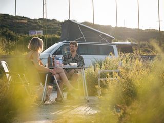 Eine Freundesgruppe sitzt beim Überwintern in Portugal um einen Campingtisch herum. Im Hintergrund steht ein Camper Van.