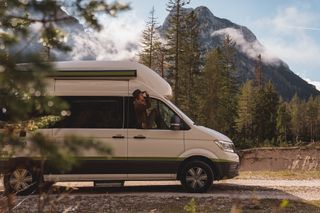 Ein Camper steht in einem Waldstück. Eine Frau sieht aus dem Fenster.