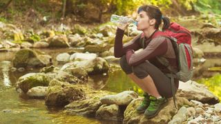 Frau beim Wandern sitz in der Hocke an einem Fluss und trinkt. 