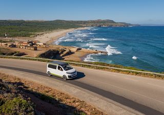 A VW California van is driving down a coastal road. The blue ocean is in the background.