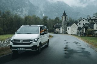 Ein Camper steht auf einer Straße. Eine Frau lehnt sich aus dem Fenster und betrachtet die Berge.