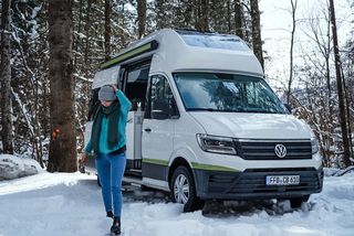 A VW Grand California is parked on a snowy forest plot. A woman in winter clothing is walking next to it.